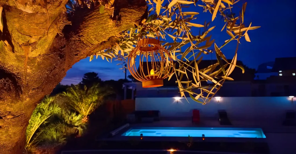 Éclairage extérieur et mise en valeur nocturne d'un jardin avec terrasse, piscine et olivier. Paysagiste Côte de Jade - Thomas Paysage